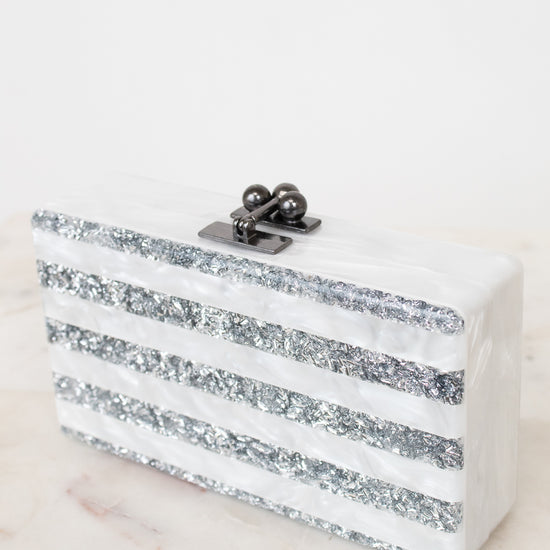 A rectangular clutch bag with horizontal white and silver glitter stripes, featuring a dark metal clasp with pearl-like accents, set on a light marble surface against a white background.