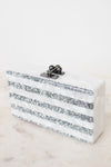 A rectangular clutch bag with horizontal white and silver glitter stripes, featuring a dark metal clasp with pearl-like accents, set on a light marble surface against a white background.