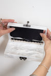 A white mother-of-pearl clutch case held open by two hands, showing a black velvet interior with a silver-tone metal hinge and clasp.