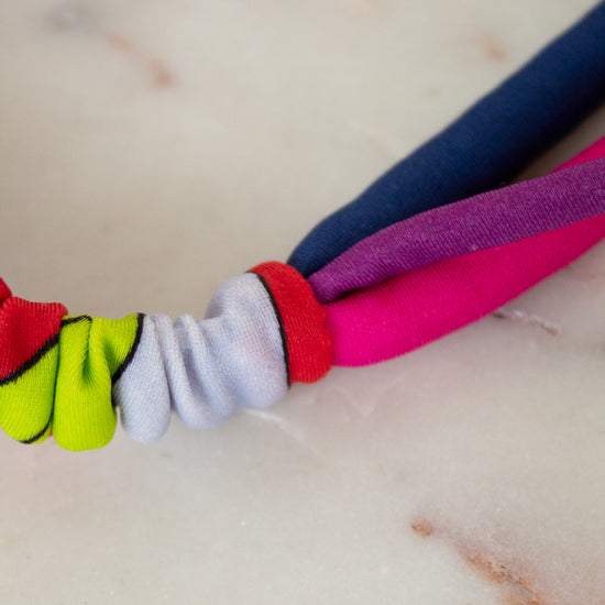 A close-up of a colorful fabric headband or hair tie with knotted fabric loops in neon yellow-green, white, red, pink, purple, and navy blue, resting on a light marble surface.