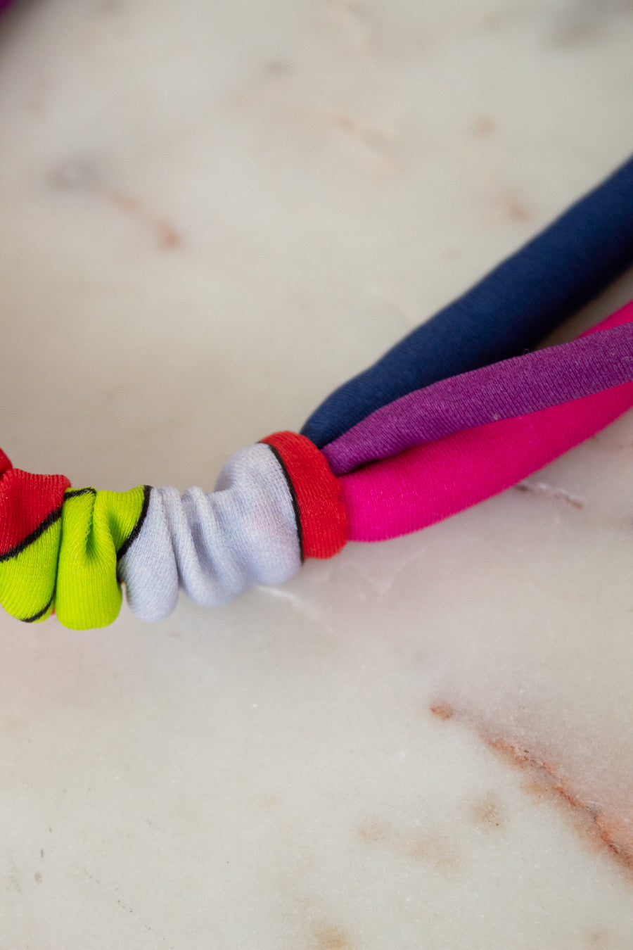 A close-up of a colorful fabric headband or hair tie with knotted fabric loops in neon yellow-green, white, red, pink, purple, and navy blue, resting on a light marble surface.
