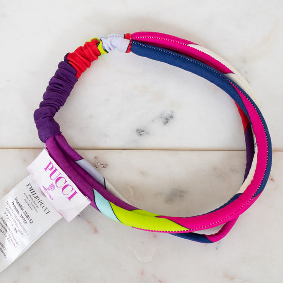 A multi-colored elastic headband by Pucci, featuring a vibrant spiral pattern of pink, purple, blue, red, and lime green, shown with its white paper tag on a white marble surface.