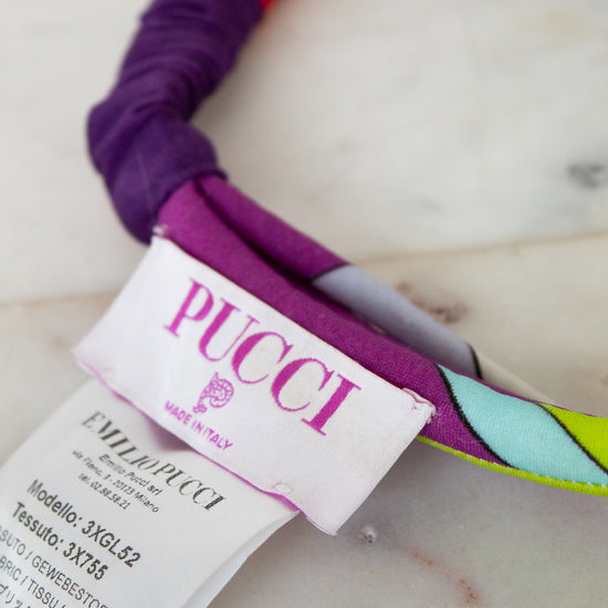 Close-up of a colorful fabric headband with a white PUCCI brand paper tag attached, resting on a light marble-like surface.