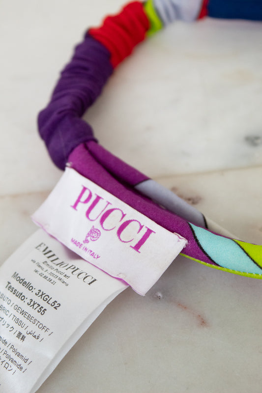 Close-up of a colorful fabric headband with a white PUCCI brand paper tag attached, resting on a light marble-like surface.