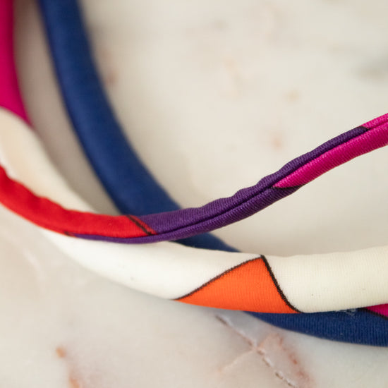 A close-up of a multi-colored fabric headband with geometric color-blocked panels in white, orange, purple, red, and navy blue, resting on a light marble surface.