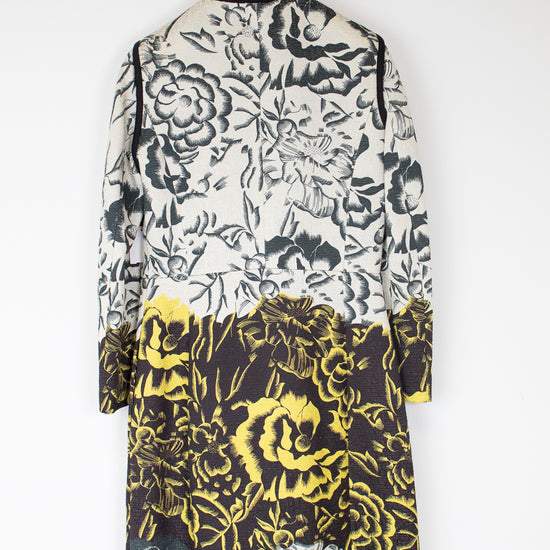 A long-sleeve floral coat hanging on a black hanger against a plain white wall. The coat features a black and white floral pattern on the upper portion and a bold yellow and black floral pattern on the lower half.