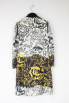 A long-sleeve floral coat hanging on a black hanger against a plain white wall. The coat features a black and white floral pattern on the upper portion and a bold yellow and black floral pattern on the lower half.