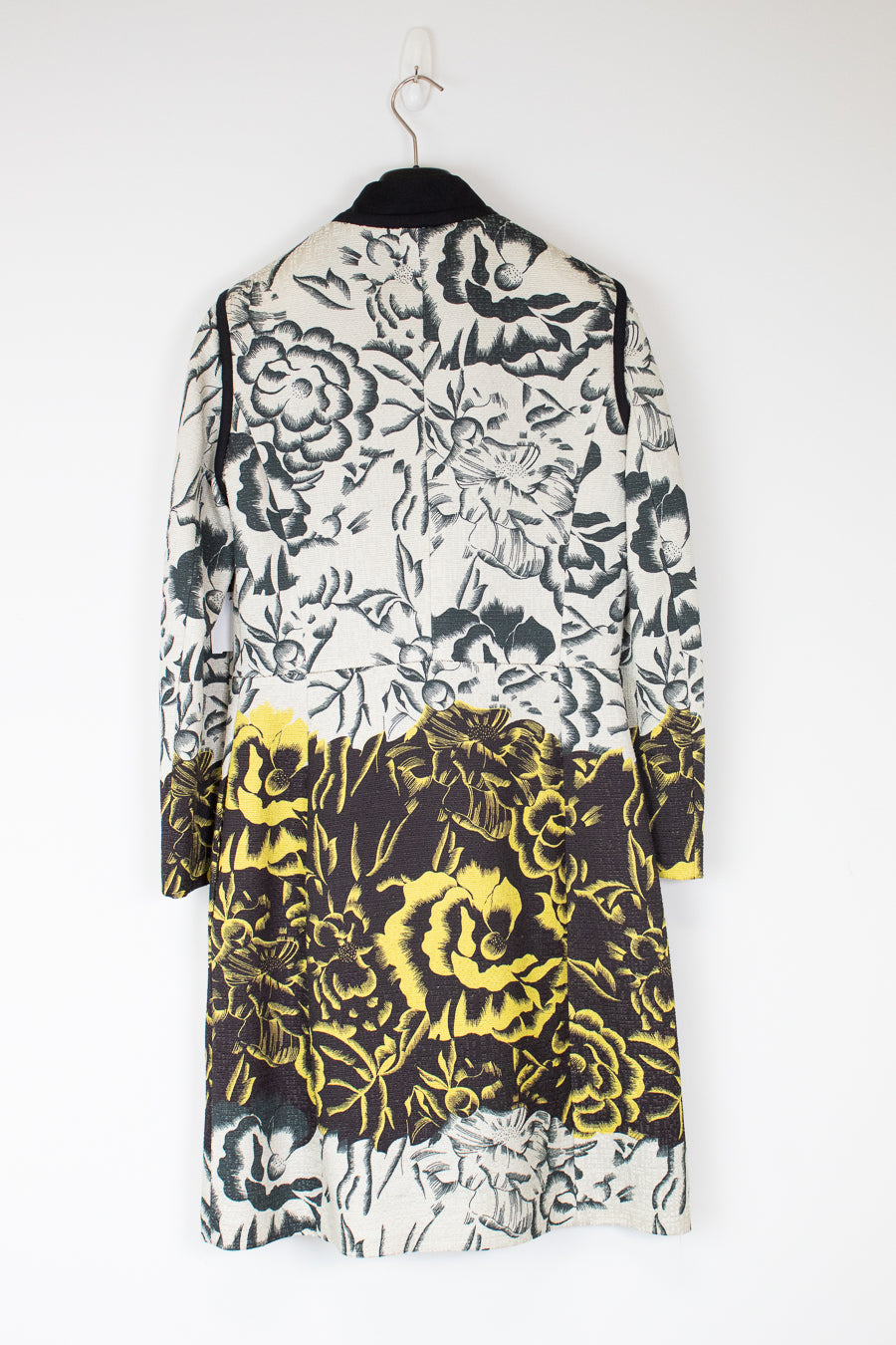 A long-sleeve floral coat hanging on a black hanger against a plain white wall. The coat features a black and white floral pattern on the upper portion and a bold yellow and black floral pattern on the lower half.