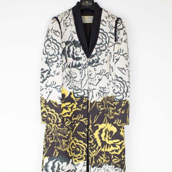 A long-sleeved floral coat with a black shawl collar, featuring a black and white floral pattern on the upper half and a black, yellow, and white floral pattern on the lower half, hanging on a white hanger against a white wall.