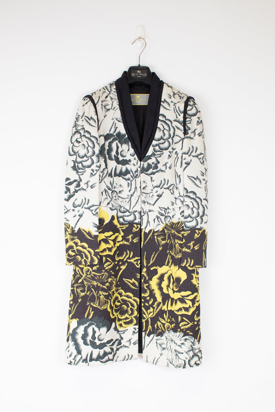 A long-sleeved floral coat with a black shawl collar, featuring a black and white floral pattern on the upper half and a black, yellow, and white floral pattern on the lower half, hanging on a white hanger against a white wall.