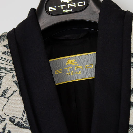 A close-up of an ETRO Milano garment hanging on a black hanger with the ETRO Milano logo. The garment has a black shawl collar and inner lining, with a patterned off-white and black outer fabric visible on the sides.