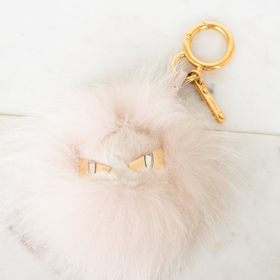 A fluffy pale pink faux-fur keychain or bag charm with small gold eye details and a gold key ring, photographed on a light marble-like surface.