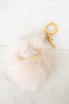 A fluffy pale pink faux-fur keychain or bag charm with small gold eye details and a gold key ring, photographed on a light marble-like surface.