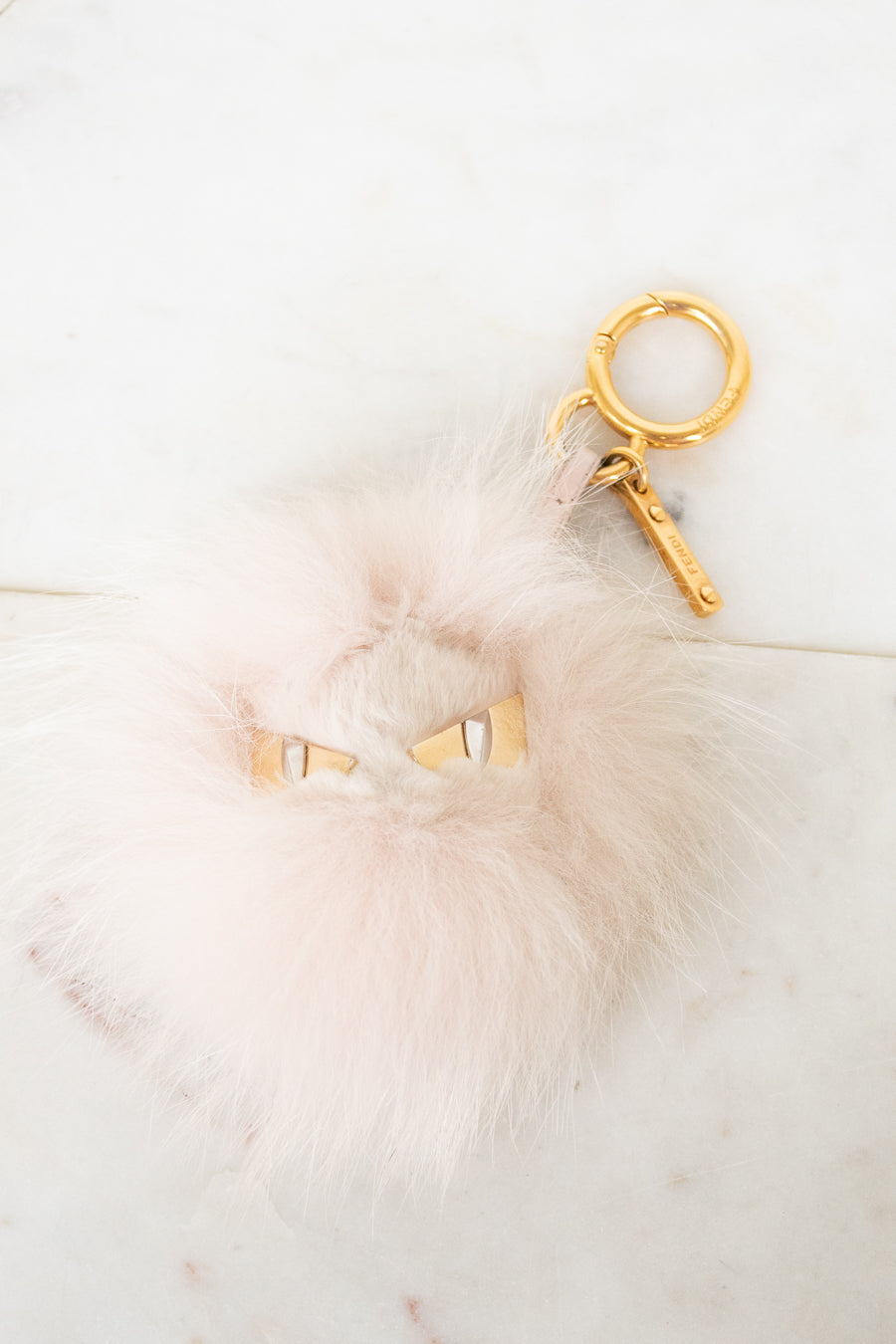 A fluffy pale pink faux-fur keychain or bag charm with small gold eye details and a gold key ring, photographed on a light marble-like surface.