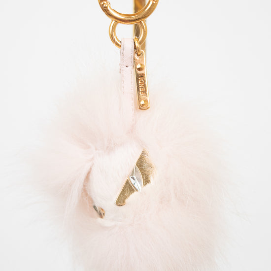 A pale pink fluffy pom-pom bag charm with gold-tone hardware, including a split key ring, keychain tag, and decorative gold foil accent, shown against a plain white background.