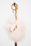 A pale pink fluffy pom-pom bag charm with gold-tone hardware, including a split key ring, keychain tag, and decorative gold foil accent, shown against a plain white background.