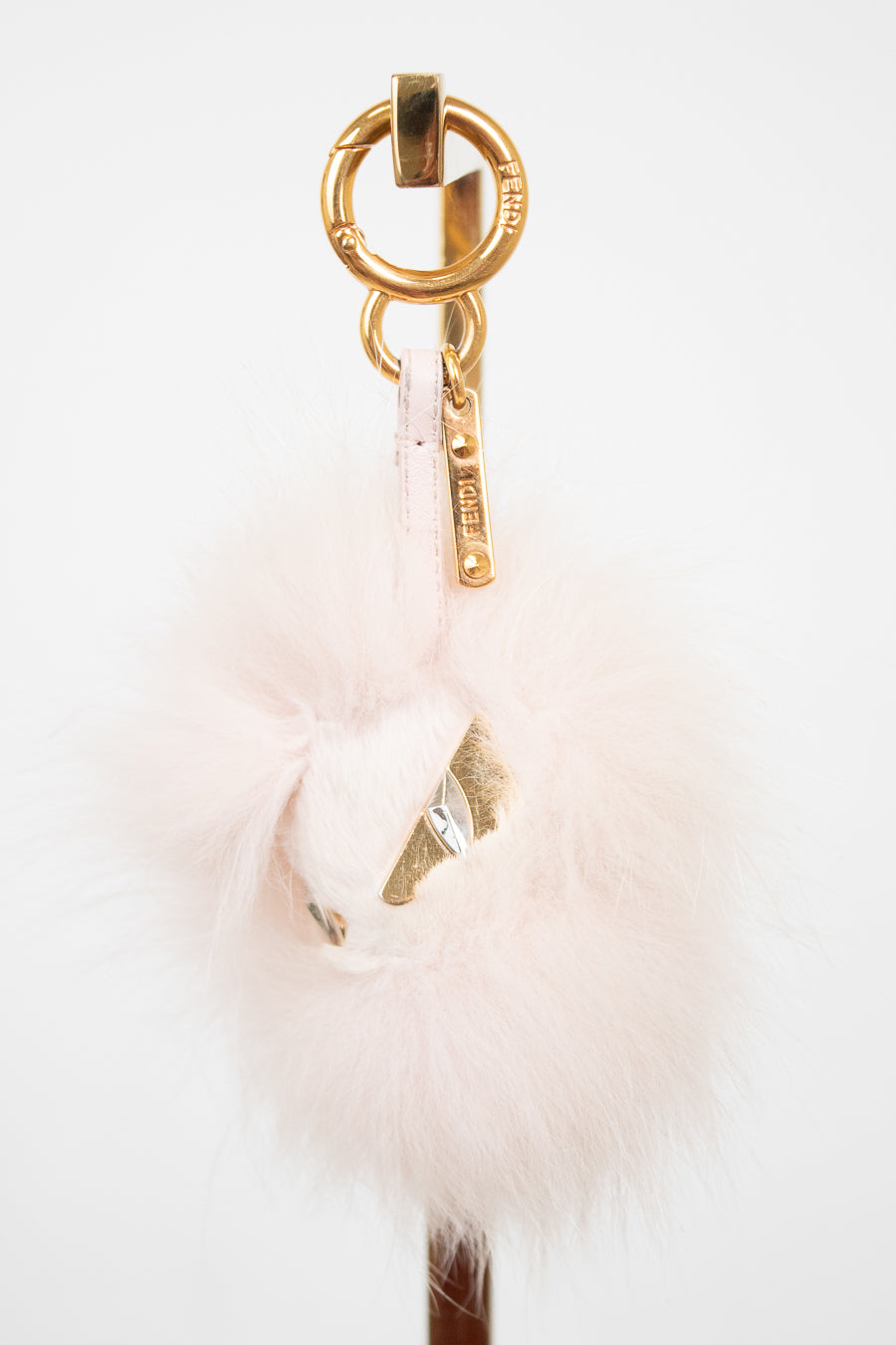 A pale pink fluffy pom-pom bag charm with gold-tone hardware, including a split key ring, keychain tag, and decorative gold foil accent, shown against a plain white background.
