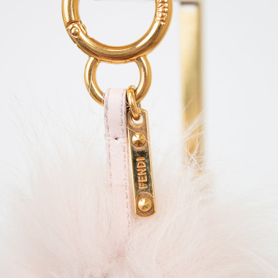 Close-up of a Fendi bag charm featuring a gold-tone metal O-ring, a gold Fendi logo tag, and a pale pink feather pom-pom on a white background.