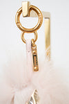 Close-up of a Fendi bag charm featuring a gold-tone metal O-ring, a gold Fendi logo tag, and a pale pink feather pom-pom on a white background.
