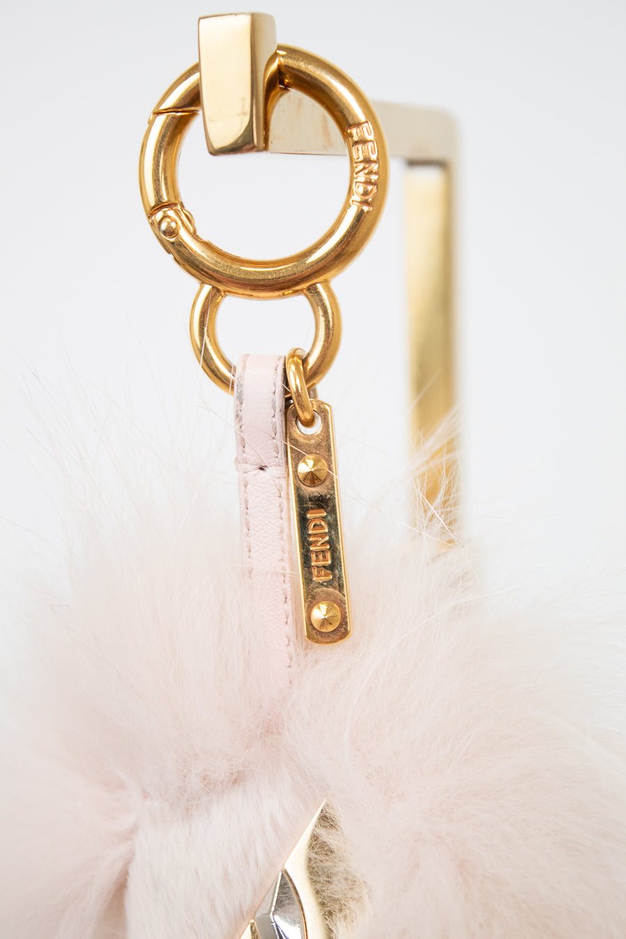 Close-up of a Fendi bag charm featuring a gold-tone metal O-ring, a gold Fendi logo tag, and a pale pink feather pom-pom on a white background.