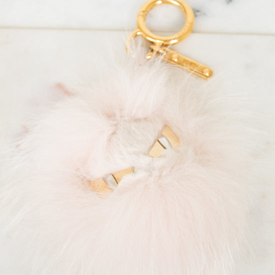 A fluffy pale pink faux-fur keychain or bag charm with gold-tone metal hardware, including a split key ring, clasp, and small decorative gold pieces, photographed on a light marble-like surface.