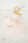 A fluffy pale pink faux-fur keychain or bag charm with gold-tone metal hardware, including a split key ring, clasp, and small decorative gold pieces, photographed on a light marble-like surface.
