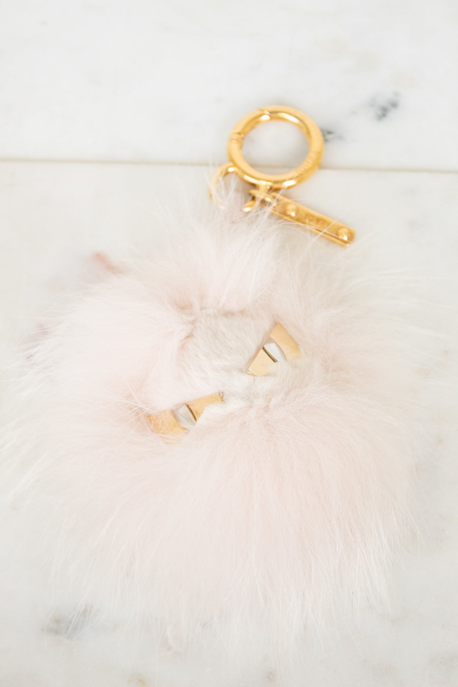 A fluffy pale pink faux-fur keychain or bag charm with gold-tone metal hardware, including a split key ring, clasp, and small decorative gold pieces, photographed on a light marble-like surface.