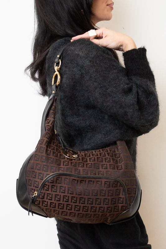 A person wearing a dark fuzzy sweater and black pants carries a brown Fendi shoulder bag with the signature FF monogram pattern and gold-tone hardware against a plain off-white background.