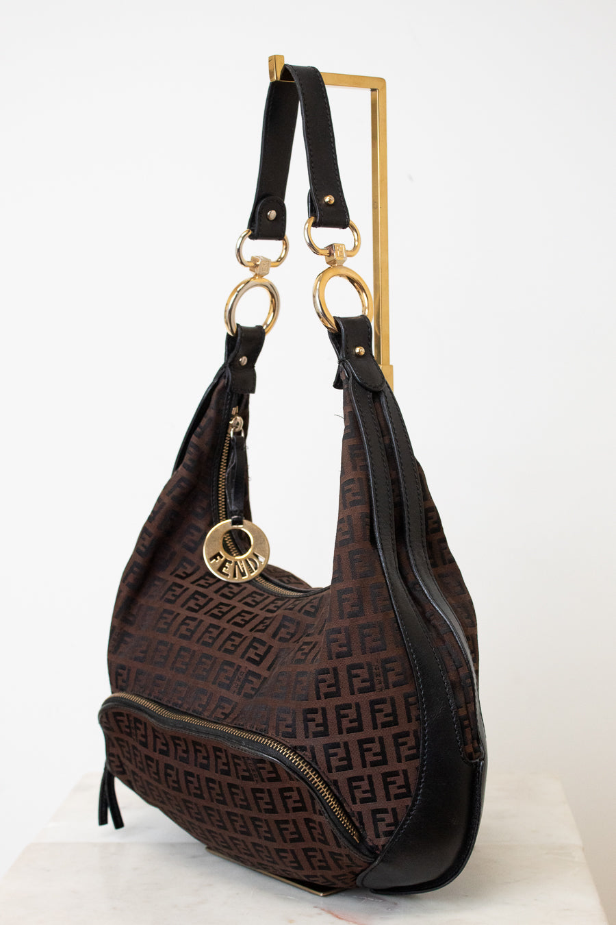 A Fendi hobo shoulder bag in the classic brown and black Zucca monogram canvas, featuring black leather trim and gold-tone hardware, including a prominent circular FF logo charm and double rings on the strap.