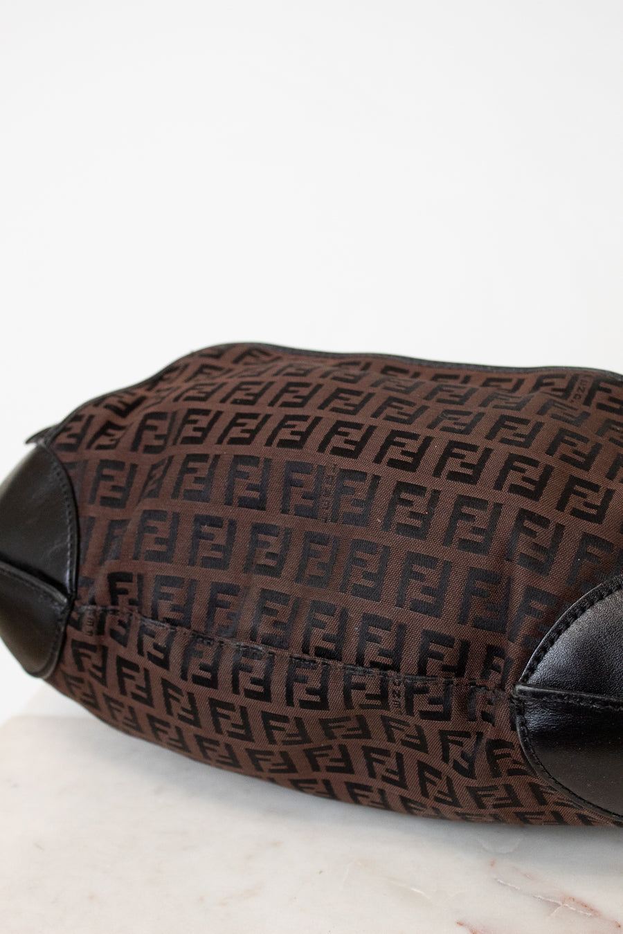 A close-up of a brown Fendi handbag featuring the signature Zucca monogram canvas and black leather trim, resting on a light marble surface against a white background.