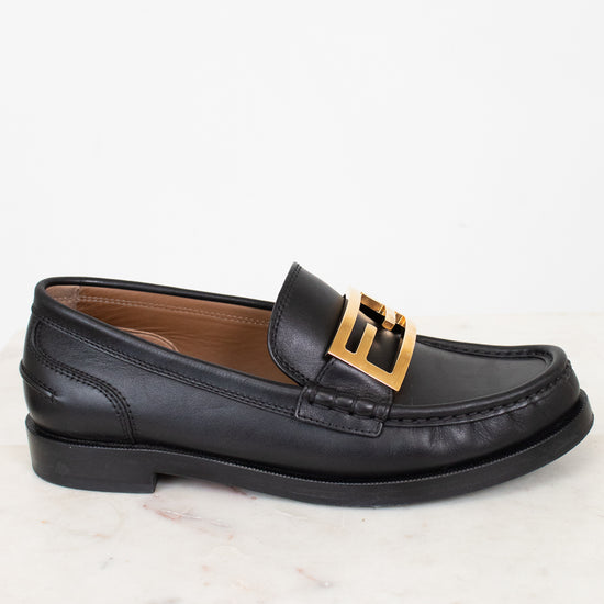 Black loafer with gold buckle on a white marble surface