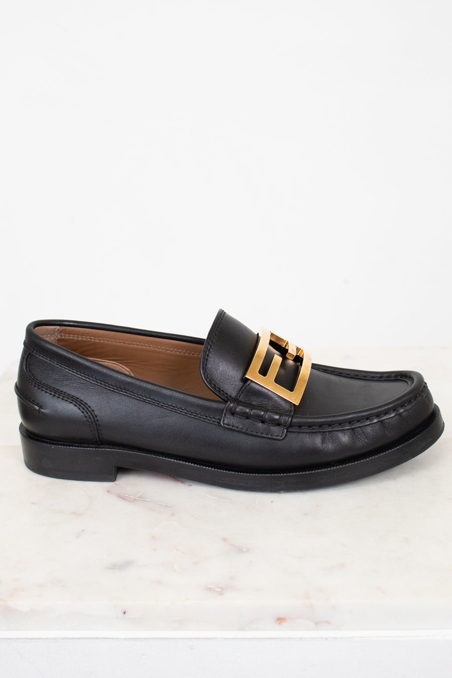 Black loafer with gold buckle on a white marble surface