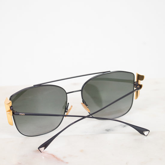 A pair of designer sunglasses with a aviator bridge design, featuring a matte black metal frame, dark lenses, and gold-tone accents on the temples, resting on a white marble surface.