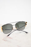 A pair of designer sunglasses with a aviator bridge design, featuring a matte black metal frame, dark lenses, and gold-tone accents on the temples, resting on a white marble surface.