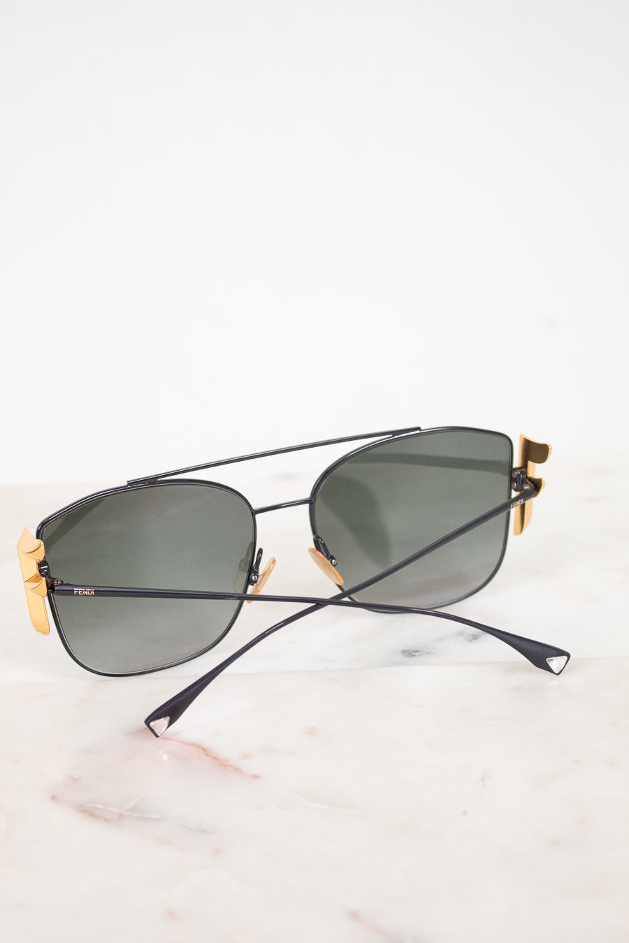 A pair of designer sunglasses with a aviator bridge design, featuring a matte black metal frame, dark lenses, and gold-tone accents on the temples, resting on a white marble surface.