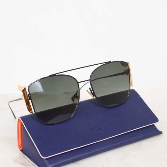 Fendi sunglasses with dark lenses and gold metal frames, resting on a navy blue Fendi-branded case against a clean white background.