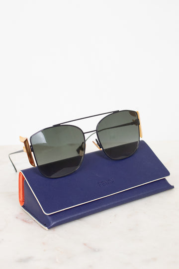Fendi sunglasses with dark lenses and gold metal frames, resting on a navy blue Fendi-branded case against a clean white background.