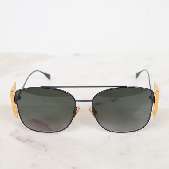 A pair of aviator-style sunglasses with dark lenses, a thin black metal frame, and distinctive gold-toned side temples, displayed on a light marble surface against a white background.