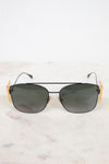 A pair of aviator-style sunglasses with dark lenses, a thin black metal frame, and distinctive gold-toned side temples, displayed on a light marble surface against a white background.