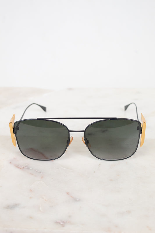 A pair of aviator-style sunglasses with dark lenses, a thin black metal frame, and distinctive gold-toned side temples, displayed on a light marble surface against a white background.