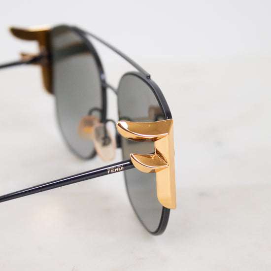 Close-up of a pair of sunglasses with dark lenses, black metal frames, and prominent gold metal hinge accents, resting on a light marble surface.