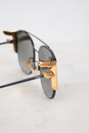 Close-up of a pair of sunglasses with dark lenses, black metal frames, and prominent gold metal hinge accents, resting on a light marble surface.