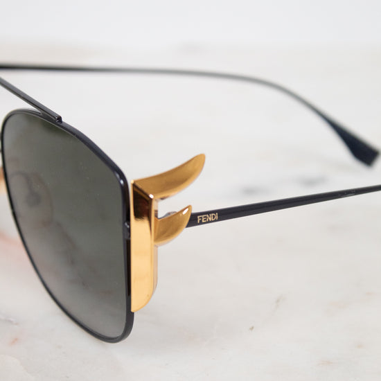 Fendi sunglasses with a black frame, dark lenses, and signature gold metal hardware, resting on a light marble surface.