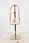 A small blush pink handbag with a thick braided shoulder strap and gold-tone hardware, displayed on a gold stand against a white studio background.