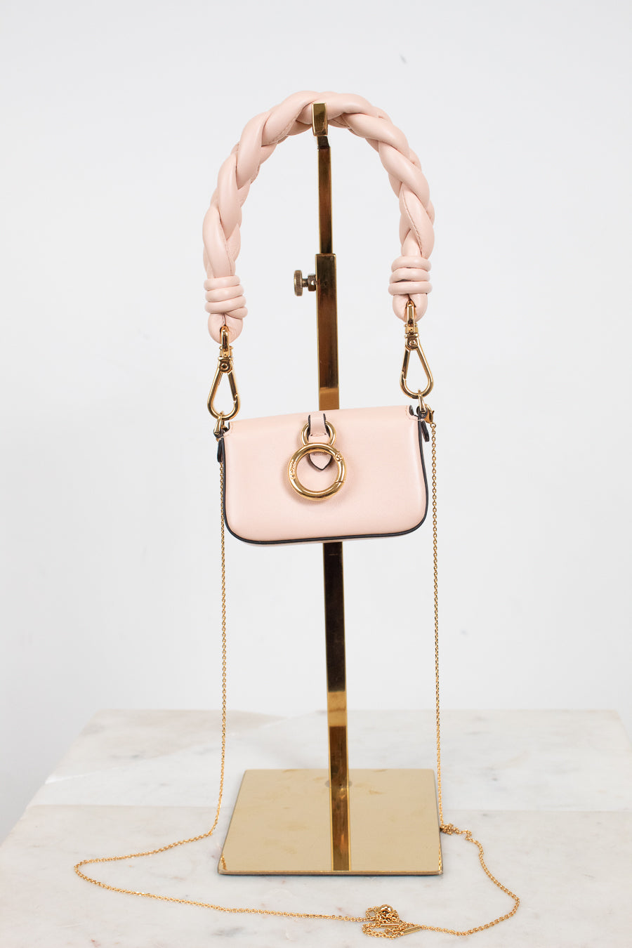A small blush pink handbag with a thick braided shoulder strap and gold-tone hardware, displayed on a gold stand against a white studio background.