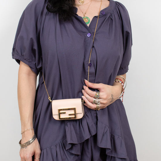 A woman wearing a short-sleeve ruffled mini dress in a muted purple/mauve color, styled with a small beige crossbody bag, layered gold necklaces, and rings, against a plain light gray background.