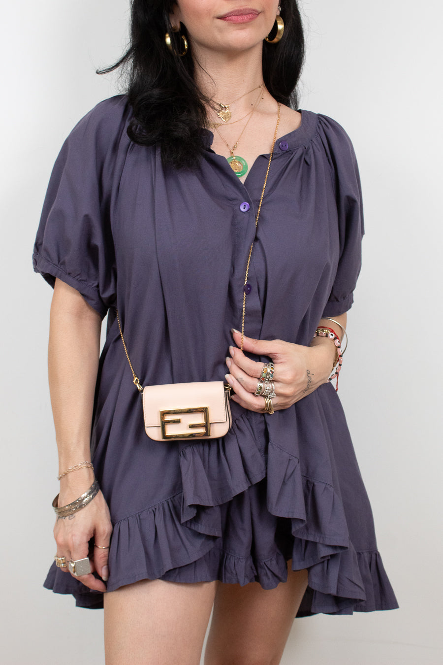 A woman wearing a short-sleeve ruffled mini dress in a muted purple/mauve color, styled with a small beige crossbody bag, layered gold necklaces, and rings, against a plain light gray background.