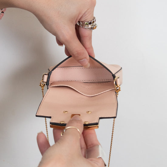 A close-up of a person's hands holding open a small structured light pink leather handbag with gold-tone hardware and a gold chain strap against a plain light background.