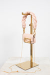 A light pink leather shoulder bag with a chunky braided shoulder strap and gold-tone hardware, displayed on a gold metal stand against a white background.