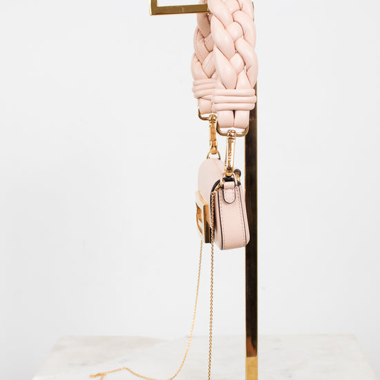 A light pink braided leather shoulder strap with gold-tone hardware, displayed on a gold metal stand next to a matching small pink handbag with a chain strap, set against a clean white background.